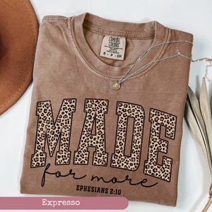 K&ouml;nnte beinhalten: Ein braunes T-Shirt mit dem Wort "MADE" in Leopardenmuster-Buchstaben und "for more" in Schreibschrift. Der Text "Epheser 2:10" ist ebenfalls zu sehen. Eine silberne Halskette mit einem kleinen Anh&auml;nger liegt auf dem Shirt.