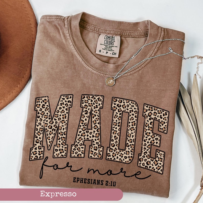 K&ouml;nnte beinhalten: Ein braunes T-Shirt mit dem Wort "MADE" in Leopardenmuster-Buchstaben und "for more" in Schreibschrift. Der Text "Epheser 2:10" ist ebenfalls zu sehen. Eine silberne Halskette mit einem kleinen Anh&auml;nger liegt auf dem Shirt.