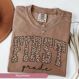 May include: A brown t-shirt with the word "FIRST" in leopard print and the words "grade" in cursive script below. The shirt is a comfortable, relaxed fit and is perfect for a first grader.