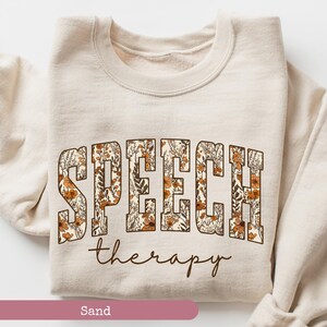May include: Sand-colored sweatshirt with the words "SPEECH" in a floral design and "therapy" in a script font. The sweatshirt is folded, showcasing the design prominently. The overall aesthetic is vintage and cozy.