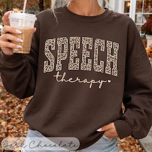 May include: A dark chocolate brown crewneck sweatshirt featuring the word SPEECH in a block font with a tan and black leopard print pattern, followed by the word therapy in a cursive script font with a small heart icon at the end.