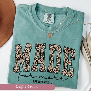K&ouml;nnte beinhalten: Hellgr&uuml;nes T-Shirt mit den Worten "MADE for more" in einem Leopardenmuster. Der Text "Ephesians 2:10" steht darunter. Eine goldene Halskette mit Anh&auml;nger befindet sich auf dem Shirt.