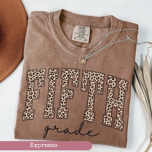 May include: A brown t-shirt with the word "FIFTH" in leopard print letters and the words "grade" in cursive script below. The shirt is labeled "Espresso".