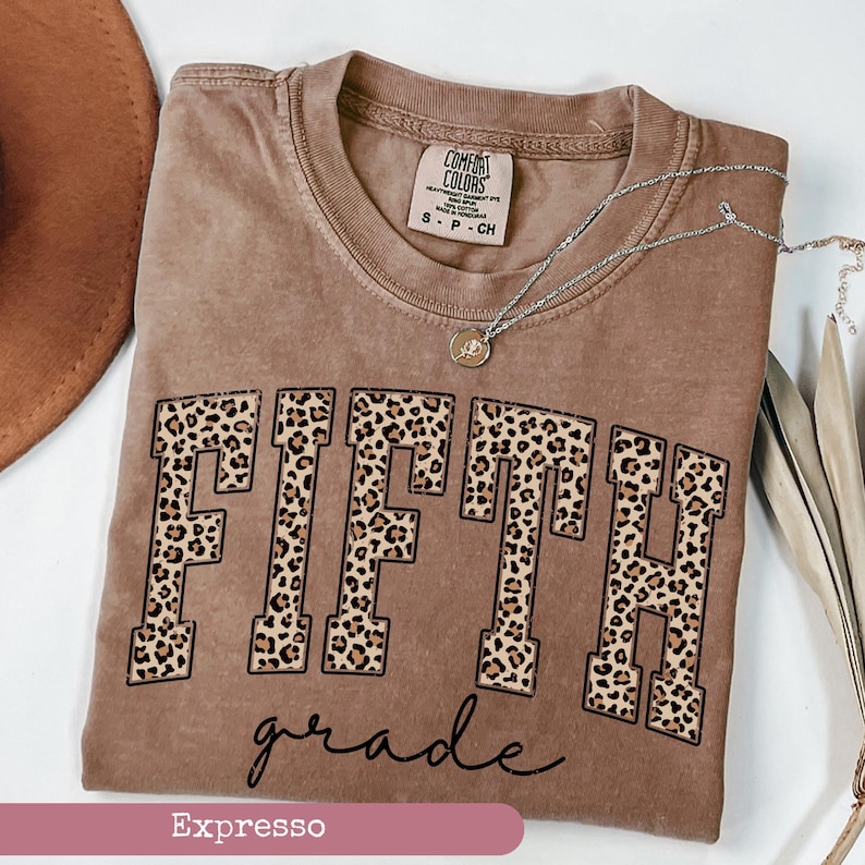 May include: A brown t-shirt with the word "FIFTH" in leopard print letters and the words "grade" in cursive script below. The shirt is labeled "Espresso".