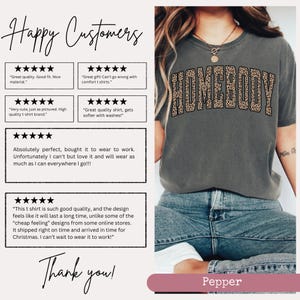 May include: A gray t-shirt with the word "HOMEBODY" in a leopard print design across the chest. The shirt is worn by a person in blue jeans. The image also includes customer reviews and the word "Pepper".