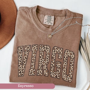 Może przedstawiać: Koszulka w kolorze espresso z nadrukiem "VIRGO" w panterkę. Koszulka ma postarzony wygląd i jest wykonana z miękkiej, wygodnej tkaniny.