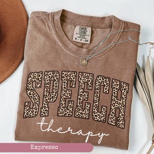May include: An espresso-colored t-shirt featuring the word SPEECH in large block letters with a leopard print pattern, followed by the word therapy in white cursive script. The shirt has a Comfort Colors brand label at the inner neckline.