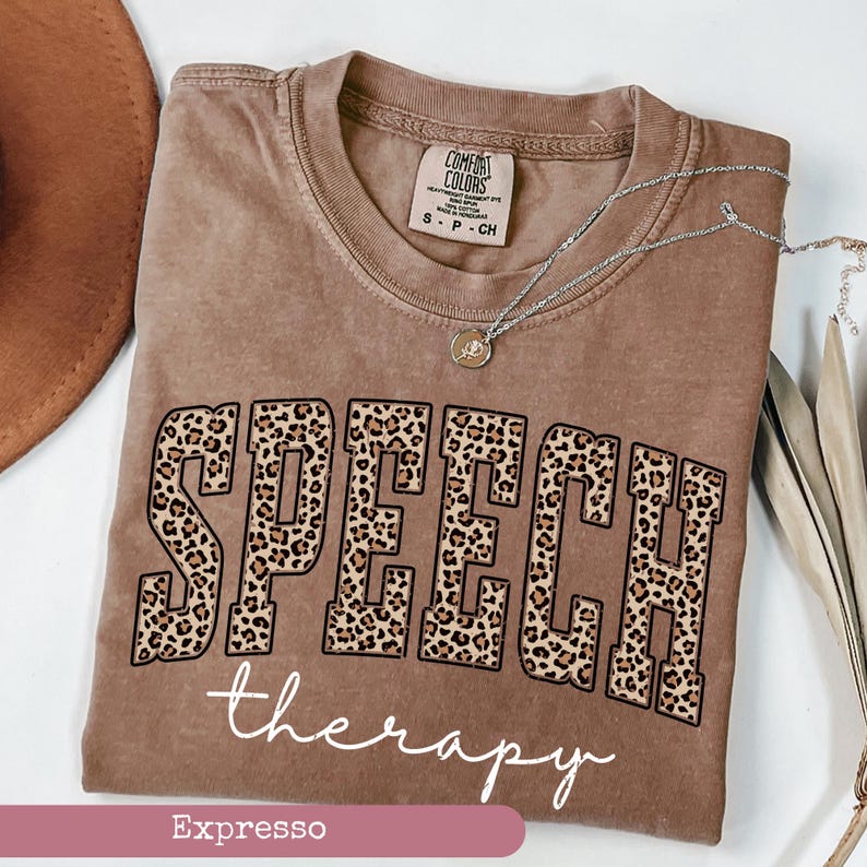May include: An espresso-colored t-shirt featuring the word SPEECH in large block letters with a leopard print pattern, followed by the word therapy in white cursive script. The shirt has a Comfort Colors brand label at the inner neckline.