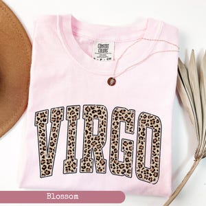 Może przedstawiać: R&oacute;żowa koszulka z nadrukiem "VIRGO" w panterkę. Koszulka jest marki Comfort Colors.