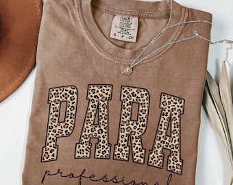 May include: A brown t-shirt with the word "PARA" in large leopard print letters, above the word "professional" in a script font. The shirt has a crew neck and short sleeves. A silver necklace with a small pendant is draped across the shirt.