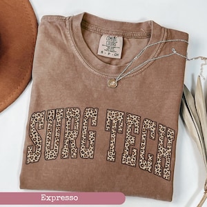 May include: A brown t-shirt with the text "SURG TECH" in a leopard print design. The shirt has a crew neck and short sleeves. A silver necklace with a small pendant is draped across the shirt.
