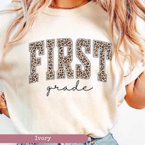 May include: A white t-shirt with a leopard print "FIRST grade" graphic. The shirt is a light ivory color.