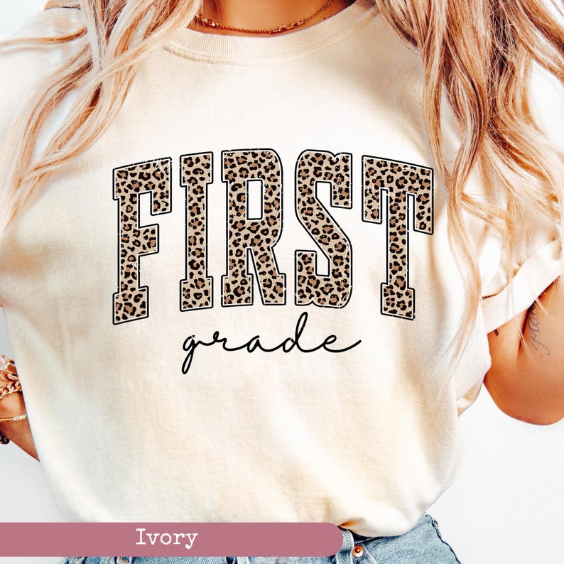 May include: A white t-shirt with a leopard print "FIRST grade" graphic. The shirt is a light ivory color.