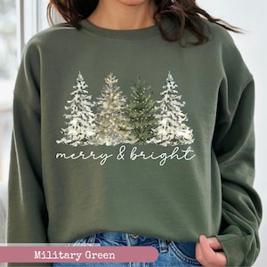 May include: Military green sweatshirt featuring a watercolor design of snow-covered Christmas trees. The phrase "merry & bright" is written in a cursive font below the trees. The sweatshirt is a solid military green color.