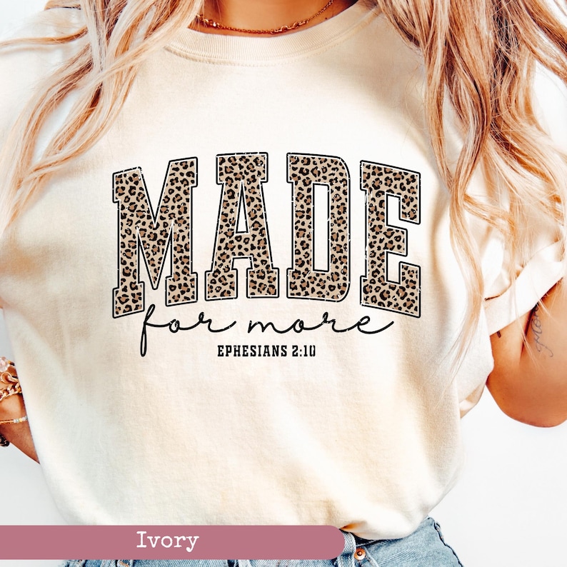 K&ouml;nnte beinhalten: Elfenbeinfarbenes T-Shirt mit dem Schriftzug "MADE for more" in einem Leopardenmuster. Der Text "Ephesians 2:10" steht darunter in einer handschriftlichen Schriftart. Das T-Shirt hat einen Rundhalsausschnitt.