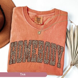 May include: A coral-colored t-shirt with the word "HOMEBODY" in large, leopard print letters. A gold necklace with a pendant rests on the shirt. The shirt is part of the Comfort Colors collection.