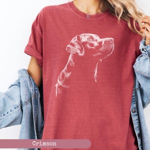 May include: A crimson red t-shirt with a white outline drawing of a Labrador Retriever dog looking to the right. The dog's head and neck are visible. The t-shirt is worn by a person with a denim jacket.