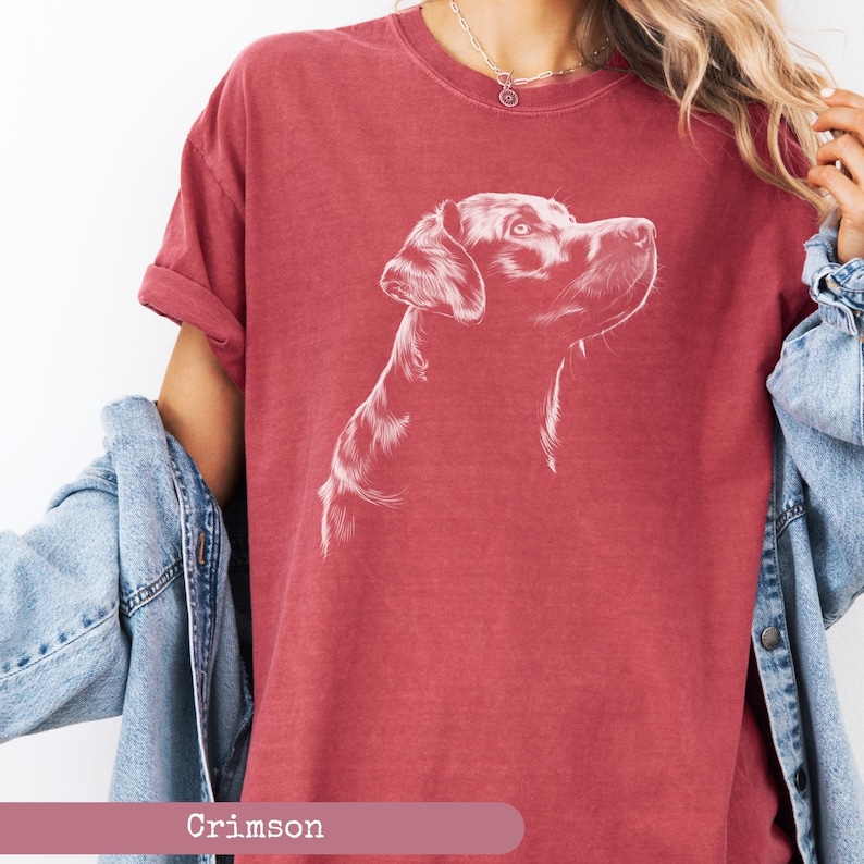 May include: A crimson red t-shirt with a white outline drawing of a Labrador Retriever dog looking to the right. The dog's head and neck are visible. The t-shirt is worn by a person with a denim jacket.