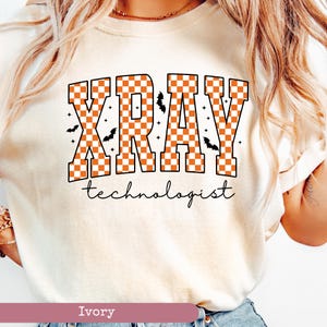 May include: Ivory-colored t-shirt with the words "XRAY technologist" in a bold, orange and white checkered design. Small black bats and stars are scattered around the text, creating a Halloween theme. The shirt is a crew neck.