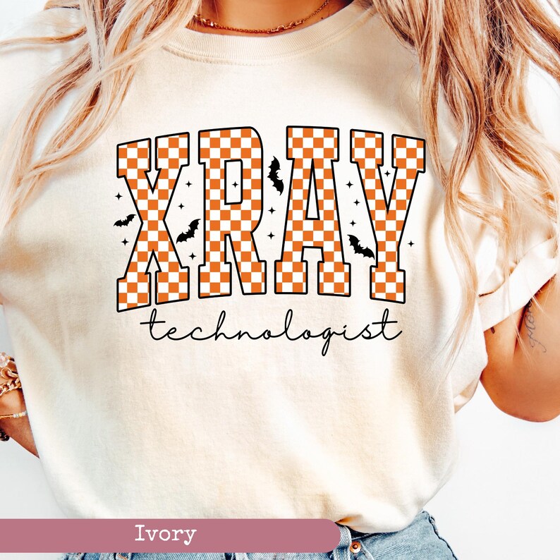 May include: Ivory-colored t-shirt with the words "XRAY technologist" in a bold, orange and white checkered design. Small black bats and stars are scattered around the text, creating a Halloween theme. The shirt is a crew neck.