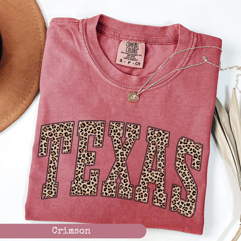 May include: Crimson-colored t-shirt with the word "TEXAS" in large, leopard print letters. The shirt has a crew neck and short sleeves. A silver necklace with a pendant is draped across the shirt. The shirt is made by Comfort Colors.