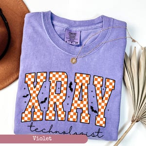 May include: A violet Comfort Colors t-shirt with the words "XRAY technologist" in orange and white checkered letters. The design includes black bats and stars. A gold necklace with a pendant is draped across the shirt.