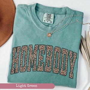 May include: A light green Comfort Colors t-shirt with the word "HOMEBODY" in a leopard print design. A gold necklace with a pendant is draped across the shirt. The shirt is folded and displayed on a white surface.