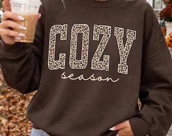 May include: A dark chocolate brown crewneck sweatshirt featuring the word COZY in large block letters with a tan and black leopard print pattern, and the word season written below in a white cursive script font across the chest area.