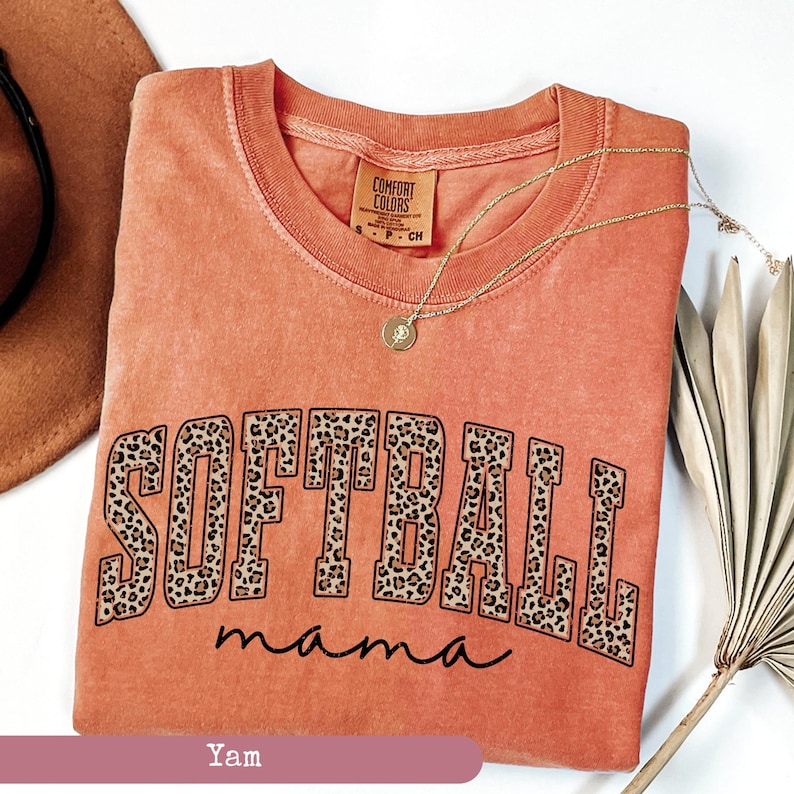 May include: A coral-colored t-shirt with the words "Softball Mama" in black lettering. The word "Softball" is outlined in brown and has a leopard print pattern.