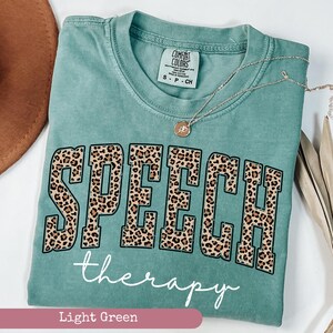 May include: A light green short-sleeve t-shirt with a crew neckline. The front features the word SPEECH in large block letters with a brown and black leopard print pattern, and the word therapy in white cursive script underneath the text.