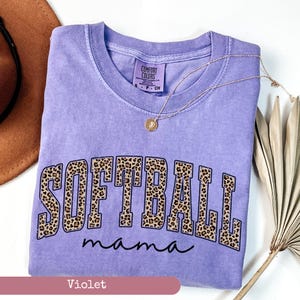 May include: A purple t-shirt with the words "SOFTBALL mama" in a leopard print design. The shirt is made by Comfort Colors.
