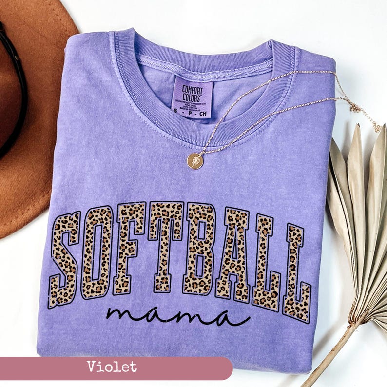 May include: A purple t-shirt with the words "SOFTBALL mama" in a leopard print design. The shirt is made by Comfort Colors.