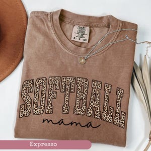 May include: A brown t-shirt with a leopard print graphic that says "Softball mama" in black text. The shirt is made by Comfort Colors and is available in sizes S, P, and CH.