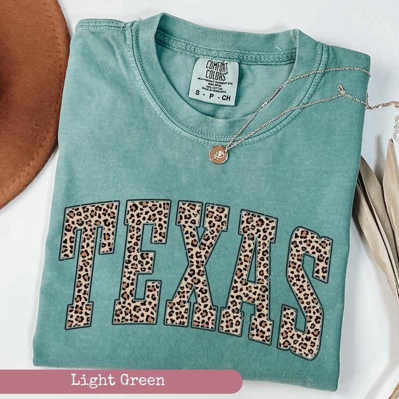 May include: A light green Comfort Colors t-shirt with the word "TEXAS" in a leopard print design. The shirt has a crew neck and short sleeves. A gold necklace with a pendant is on the shirt. The shirt is labeled "Light Green".