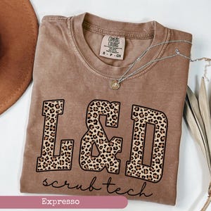 May include: A brown Comfort Colors t-shirt with the text "L&D scrub tech" in a black font with leopard print letters. A silver necklace with a small pendant is on the shirt. The shirt is size S-P-CH.