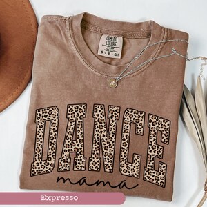 May include: A brown t-shirt with the word "DANCE" in leopard print letters. The text "mama" is written in cursive below the word "DANCE". The shirt is folded and laid flat on a surface. The word "Espresso" is written in pink at the bottom of the image.