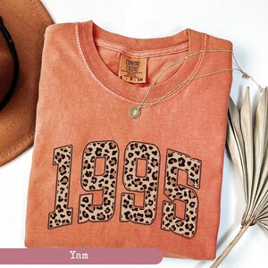 Op de afbeelding: Een koraalkleurig T-shirt met het jaar 1995 gedrukt in grote, gedurfde, zwarte letters. Elk cijfer is gevuld met een luipaardprint. Het shirt heeft een label met de tekst "Comfort Colors".