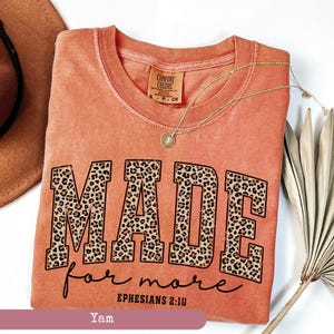 K&ouml;nnte beinhalten: Ein korallenfarbenes T-Shirt mit den Worten "MADE for more" in einer kr&auml;ftigen Leopardenmuster-Schriftart. Der Text "Ephesians 2:10" steht darunter. Eine goldene Halskette mit Anh&auml;nger liegt auf dem Shirt.