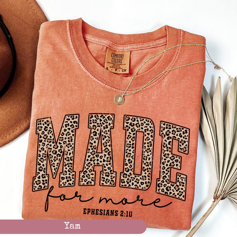 K&ouml;nnte beinhalten: Ein korallenfarbenes T-Shirt mit den Worten "MADE for more" in einer kr&auml;ftigen Leopardenmuster-Schriftart. Der Text "Ephesians 2:10" steht darunter. Eine goldene Halskette mit Anh&auml;nger liegt auf dem Shirt.