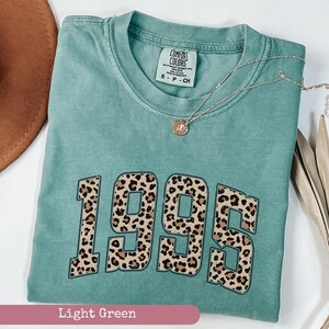 Op de afbeelding: Een lichtgroen T-shirt met een luipaardprint in de vorm van het jaar 1995. Het shirt heeft een ronde hals en korte mouwen. De tekst "COMFORT COLORS" is zichtbaar op het label.