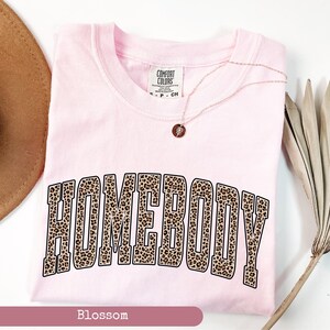 May include: A light pink t-shirt with the word "HOMEBODY" in a leopard print design. The shirt has a crew neck and short sleeves. A gold necklace with a small pendant is draped across the shirt. The shirt is labeled "Blossom" at the bottom.