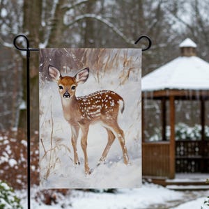 May include: A garden flag featuring a painting of a young deer, or fawn, standing in a snowy landscape. The deer has brown fur with white spots and is set against a backdrop of winter foliage and snow. The flag is suspended from a black metal hanger.