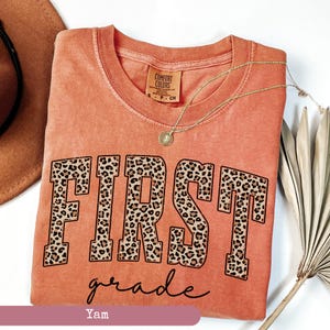 May include: A coral orange t-shirt with the word "FIRST" in black outlined letters with a leopard print pattern. The word "grade" is written in cursive below the word "FIRST".