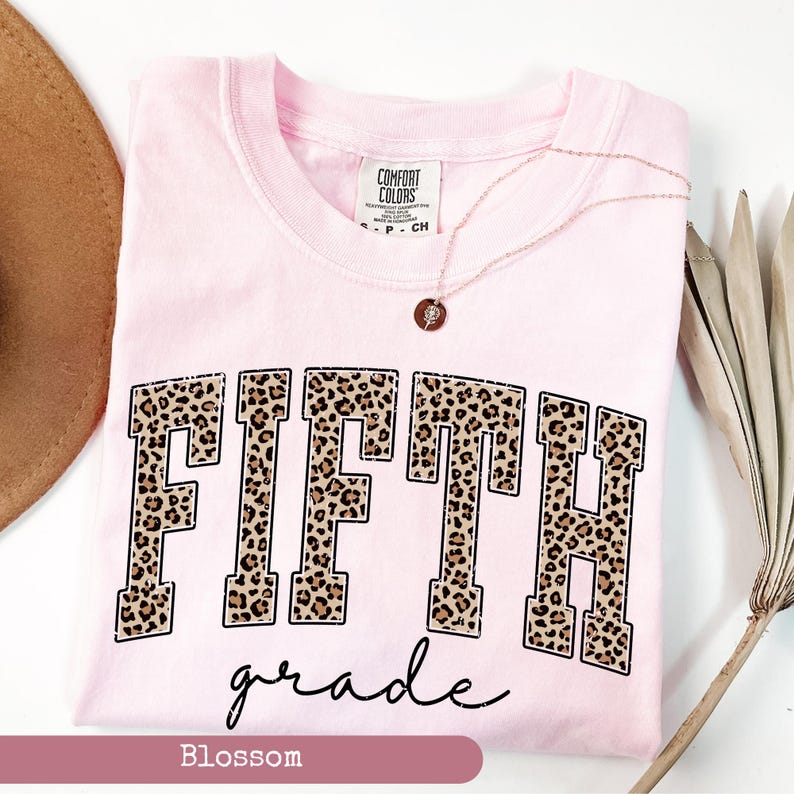 May include: Pink t-shirt with a leopard print "FIFTH grade" graphic. The shirt is a light pink color and has a distressed look. The text is in black and the leopard print is in brown and black.