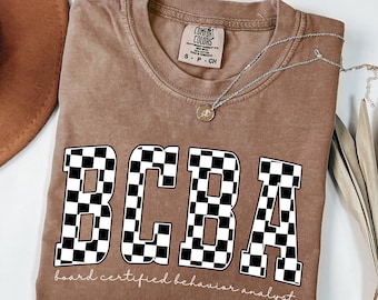 May include: Espresso-colored t-shirt with the letters "BCBA" in a bold, checkered pattern. The words "board certified behavior analyst" are printed below the letters. A silver necklace with a pendant is draped on the shirt.