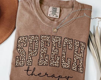 May include: A brown short-sleeved t-shirt with a crew neckline. The shirt features the word SPEECH in large block letters with a leopard print pattern, and the word therapy in black cursive script underneath. A silver necklace rests on the shirt.