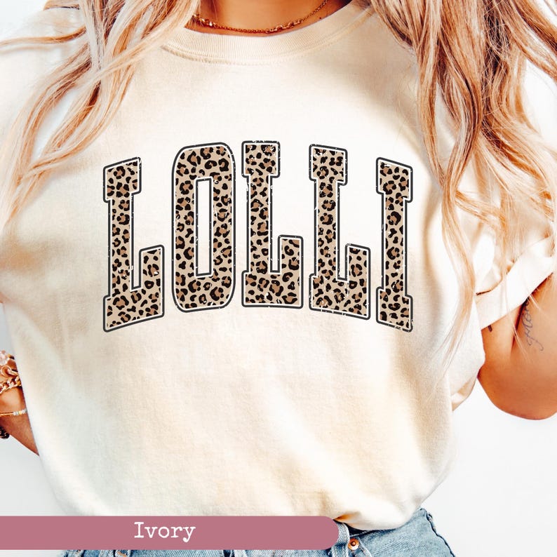 May include: A white t-shirt with the word "LOLLI" printed in a leopard print pattern. The text is in a black outline and has a vintage collegiate style font. The shirt is ivory in color.