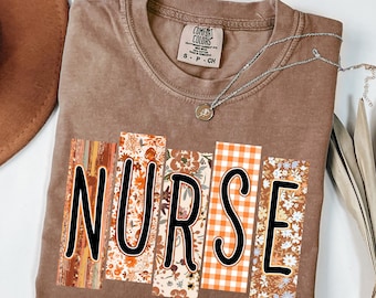 May include: Espresso-colored t-shirt with the word "NURSE" in black block letters. Each letter is filled with a different floral or plaid pattern in shades of orange, brown, and white. A silver necklace with a small pendant rests on the shirt.