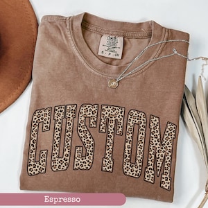 May include: Espresso-colored t-shirt with the word "CUSTOM" in large, leopard print letters. The shirt has a crew neck and short sleeves. A silver necklace with a small pendant is draped across the shirt.