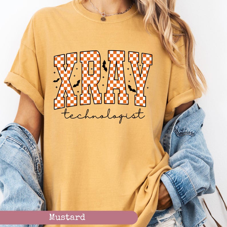 May include: Mustard-colored t-shirt with the text "XRAY technologist" in a checkered orange and white design, with small bat graphics. The shirt has rolled-up sleeves and a relaxed fit.